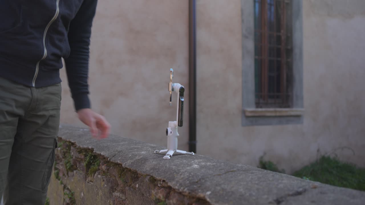 남성 관광객이 배 ⁇ 을 착용하여 이동식 짐벌에 삼각대 다리를  ⁇ 고 낮은 돌 벽에 놓습니다.