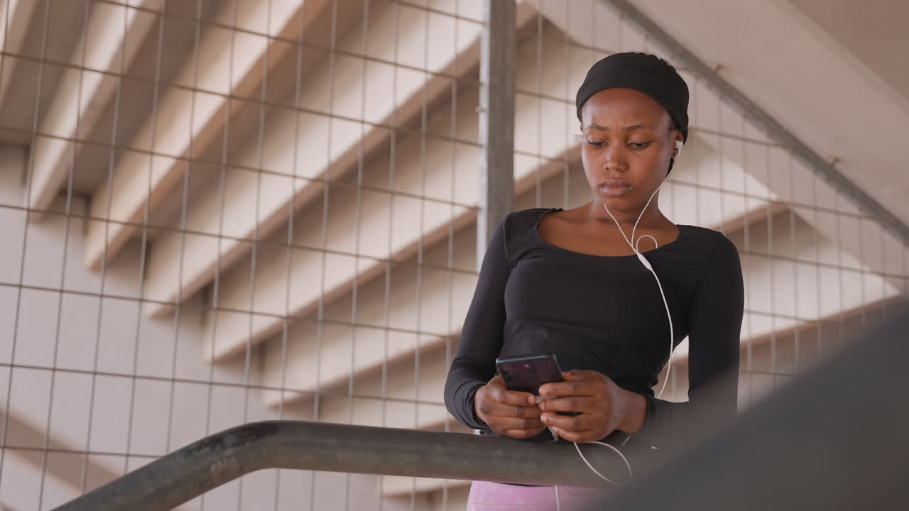 donna di colore che guarda in basso scorrendo il feed del telefono, auricolari inseriti, scala geometrica dietro, energia creativa concentrata mentre pianifica post e didascalie, momento spontaneo e tranquillo con luce naturale soffusa