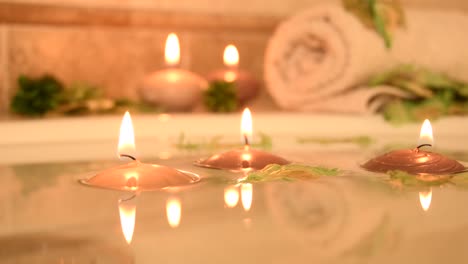 relaxing-spa-background-with-candles-floating-in-the-bath-water,-some-green-petals-and-a-towel-near-the-water-surface