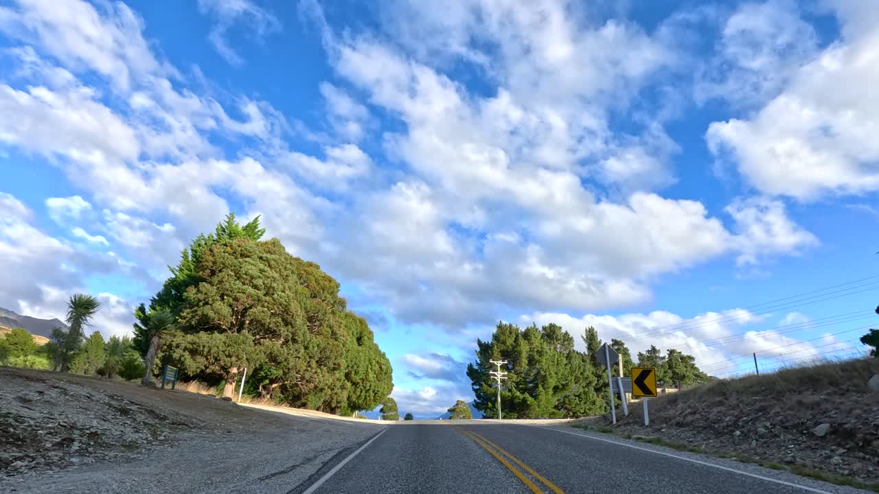 Vehicle travels rural mountain road, passing trees and farmland under bright daylight, wide-angle perspective