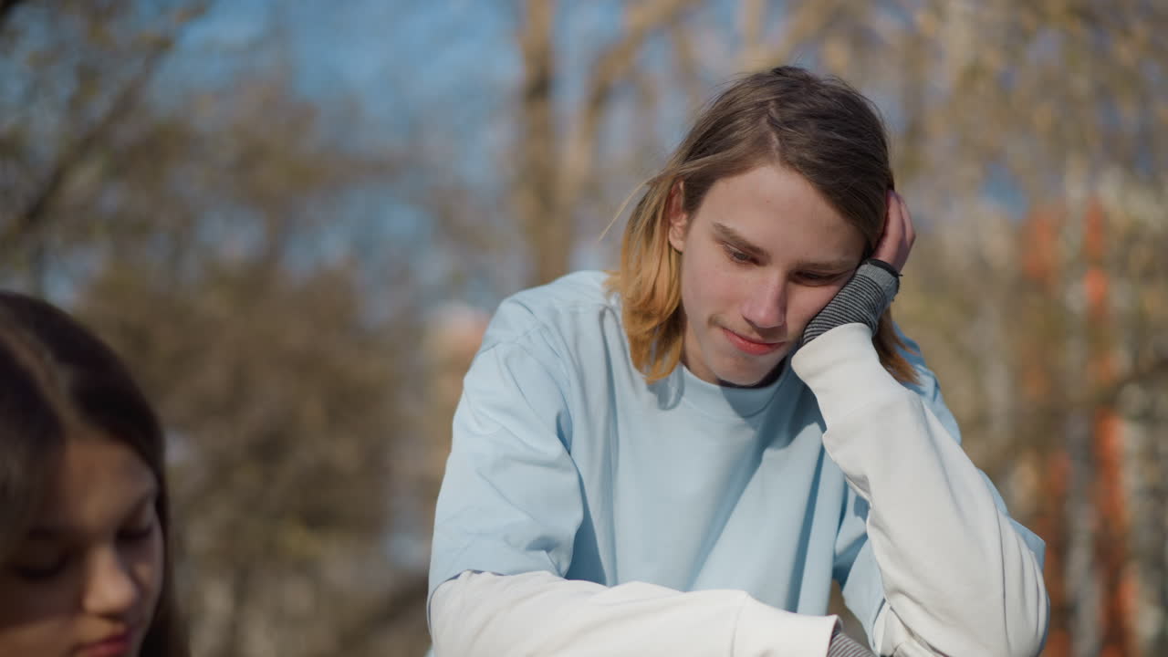 Молодой человек на улице, юноша задумчиво смотрит вдаль, подросток, сидящий в парке, выглядит погруженным в свои мысли, молодой человек с задумчивым выражением лица опирается на скамейку среди размытых деревьев в парке