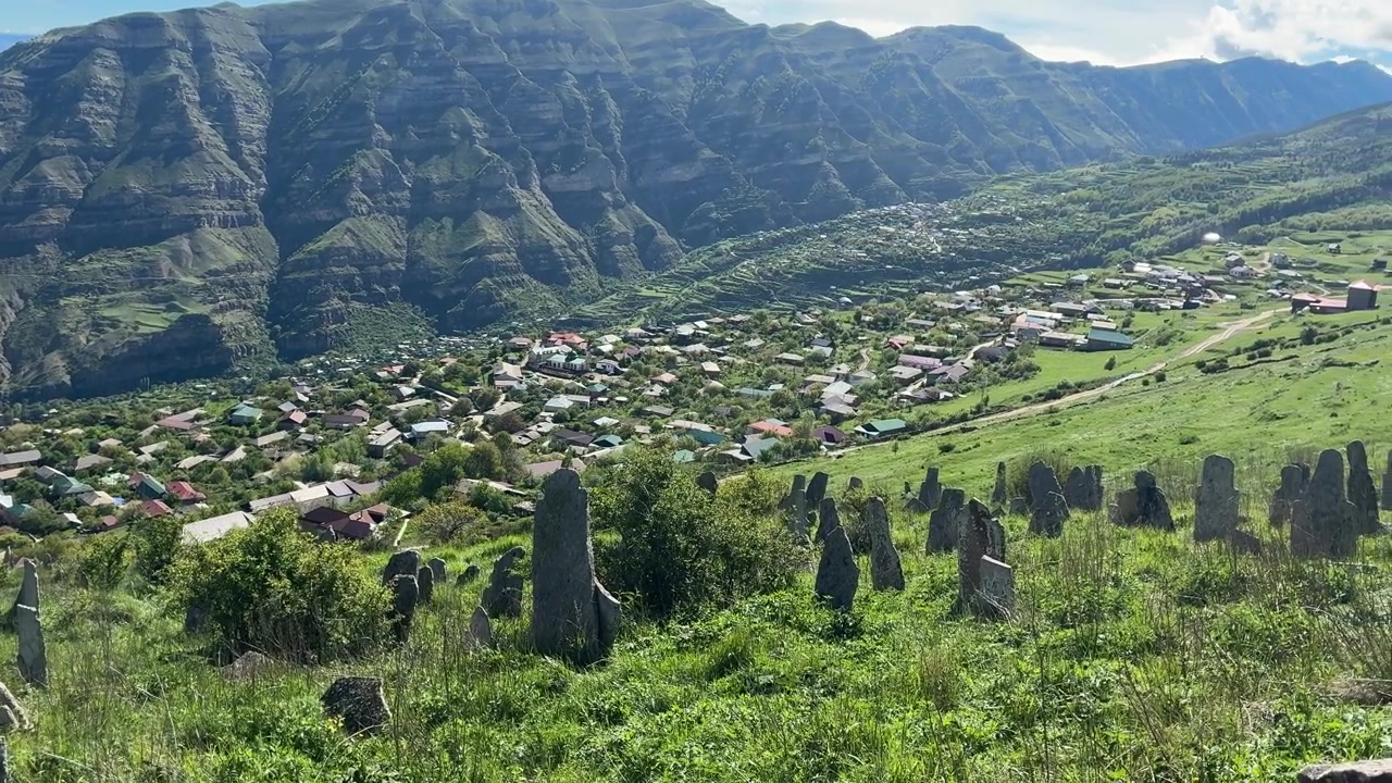 Un antico cimitero con lapidi consumate dal tempo si affaccia su un villaggio tradizionale adagiato in una valle circondata da maestose montagne nel Dagestan, Russia