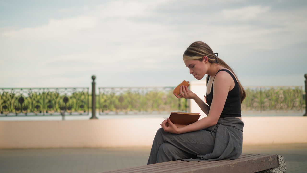 흑색 크 을 입은 여성이 야외에 앉아서, 배경에 울타리가 있는 책을 읽는 동안 햄버거를 먹고, 밝은 하늘 아래의 야외 환경에서, 캐주얼 스과 독서를 즐기고 있습니다.