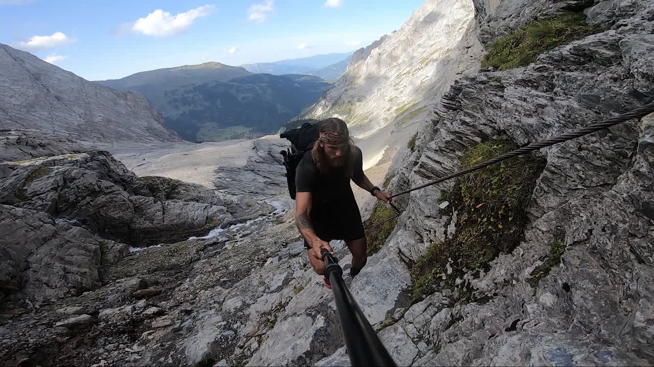 un hombre joven, fuerte y en forma, con cabello largo y tatuajes, está escalando una montaña mientras se aferra a una cuerda para ayudarlo a subir