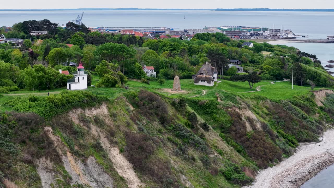 Drone footage of lush green hills and steep coastal cliffs beside the sea, featuring small houses and a lighthouse near Ega and Mols Bjerge in Denmark