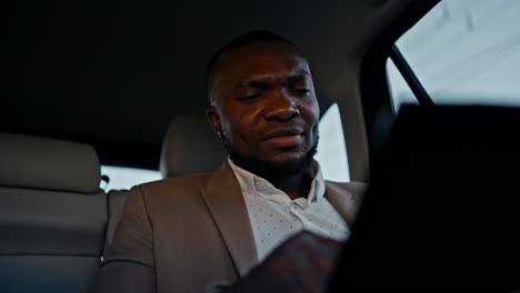 confident and thoughtful man businessman with black skin color in a brown suit reviews his plans and draws with his finger on a tablet while driving in a modern salon in a car in an urban area