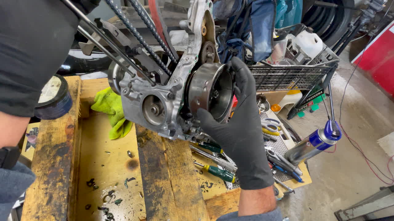 Primo piano dall'alto di un meccanico che indossa guanti neri, tiene in mano il motore di uno scooter e fa girare il volano per osservare il movimento della catena di distribuzione su un banco di legno in un garage domestico