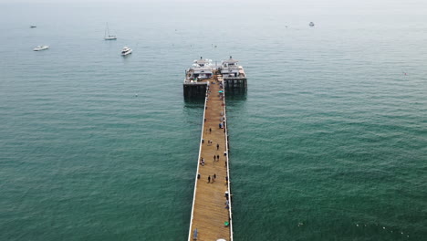 people walking along the length of malibu pier with malibu farm cafe at the end of the pier and boats in the background in california usa - descending drone shot