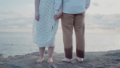 -Couple-holding-hands,-standing-barefoot-on-rocks-by-the-sea-during-sunset