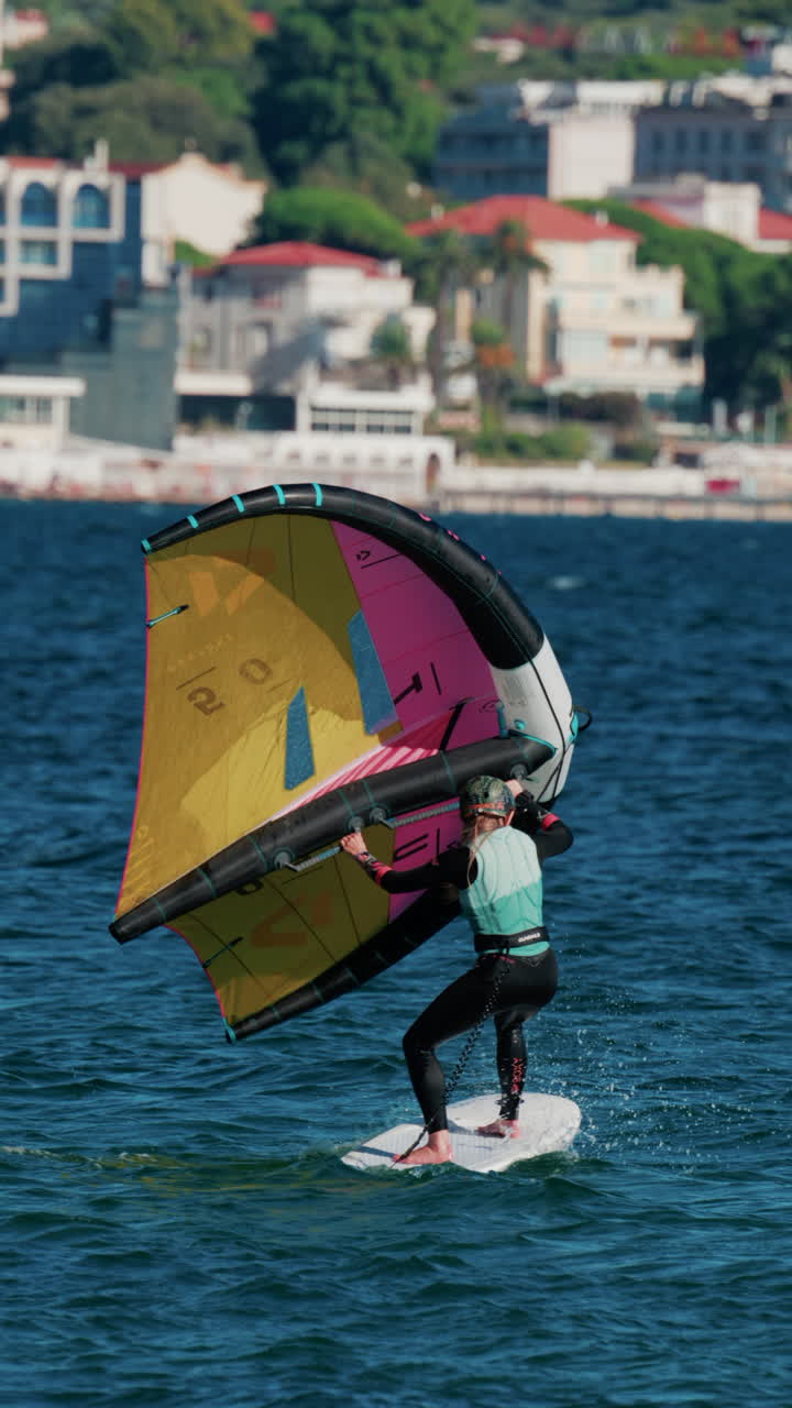 Cannes, France - October 6, 2025: A woman balances on a wing foil board holding a colorful yellow and pink sail on a sunny day. Vertical