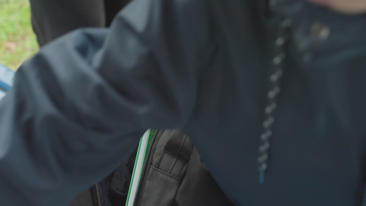 Student placing folder into backpack on bench, closeup hands zipping bag, laptop and notebooks visible, green grass background, casual jacket, morning routine before school, focus on organization