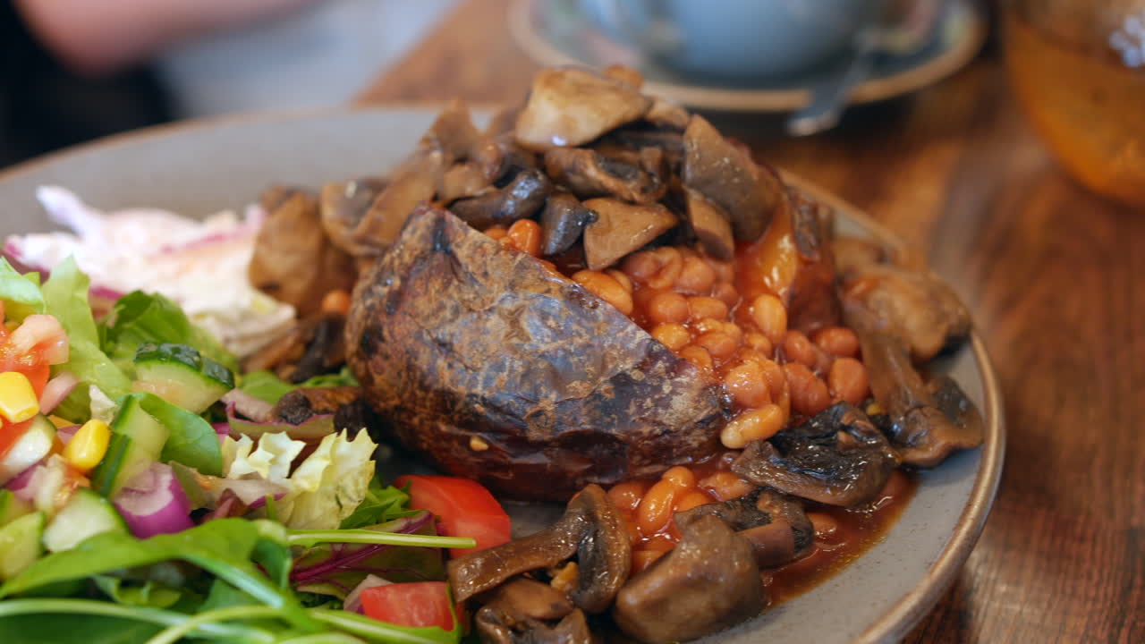 A jacket potato topped with baked beans and sautéed mushrooms sits alongside a fresh salad of mixed greens, cucumber, tomato, and corn. The dish with hearty elements with a vibrant, colorful side