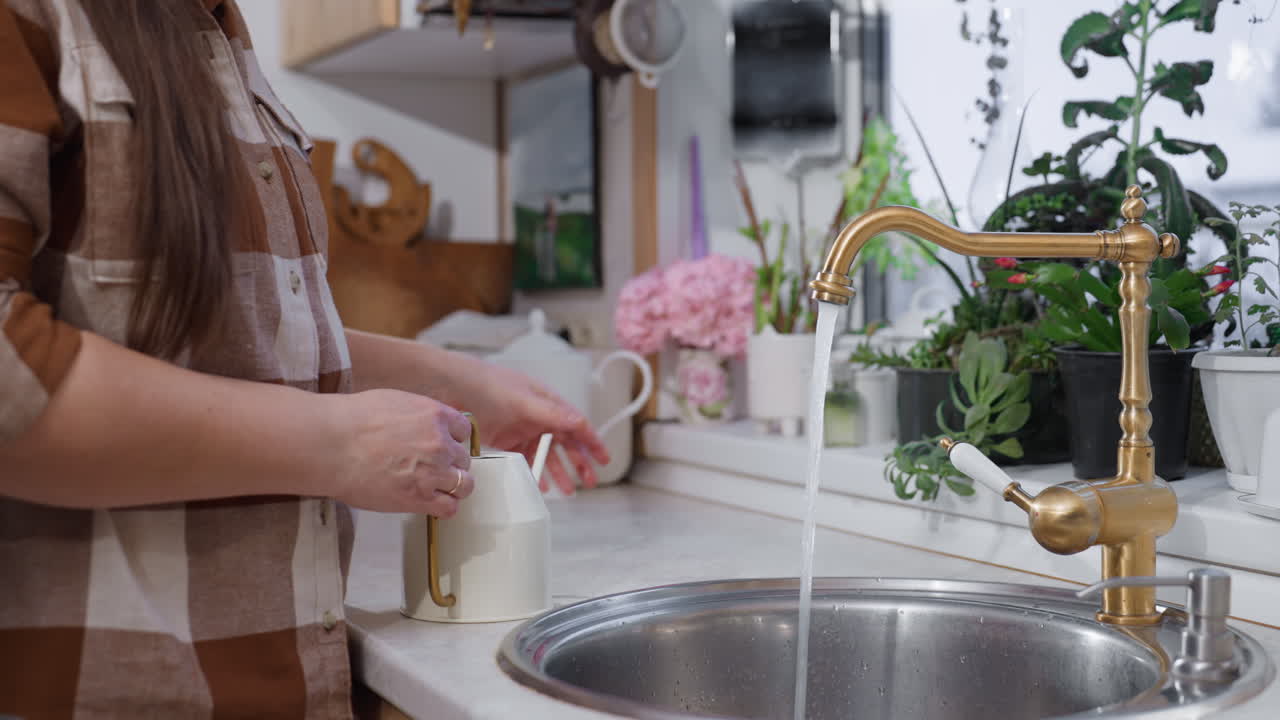 Plant lover places white kettle under running tap to fill water beside potted greenery on cozy kitchen sink ledge with brass faucet and plaid gardener in calm indoor gardening moment