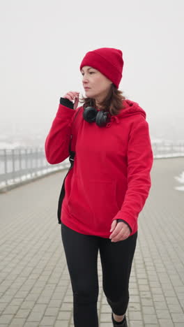athlete wearing red jacket walking along interlocked pathway with bag over shoulder, gazing away thoughtfully near iron railing and blurred city view in foggy winter atmosphere