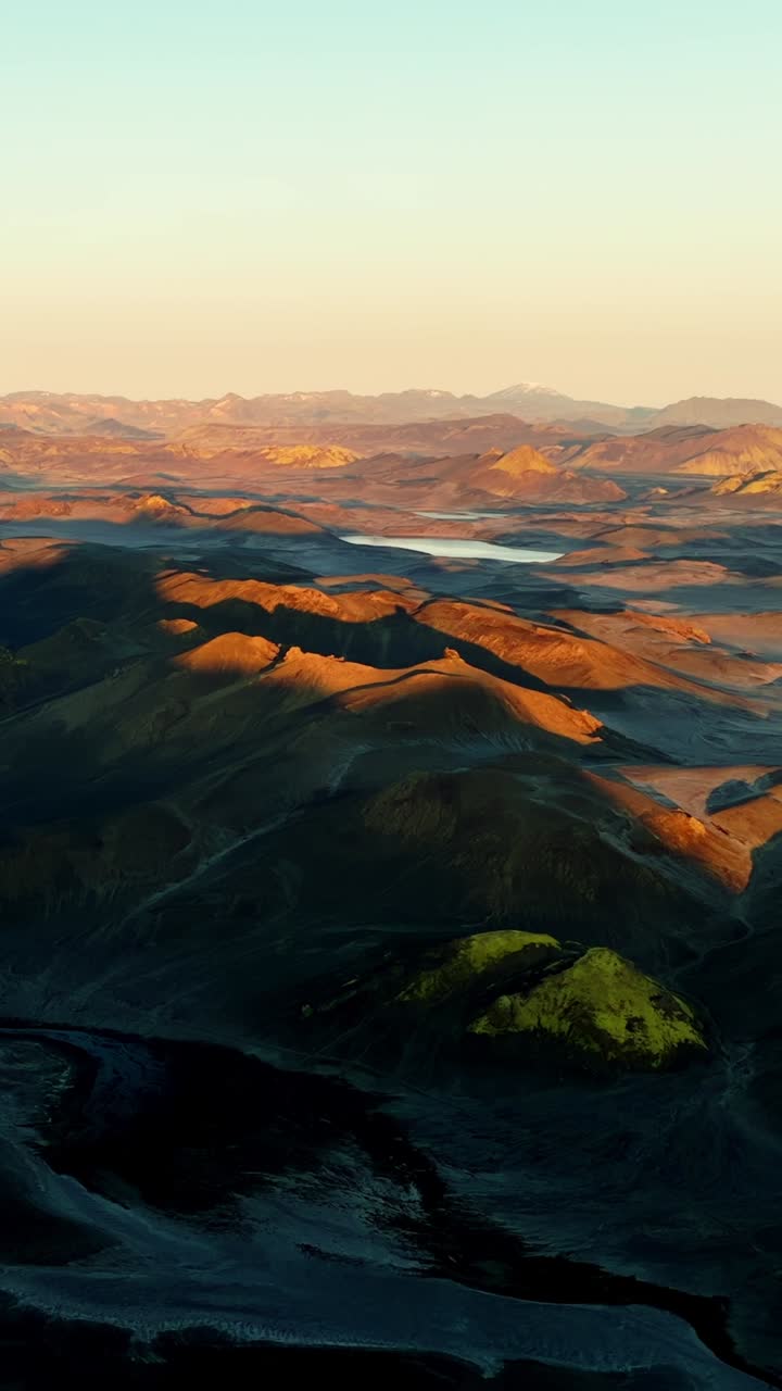 Drönarpanorama under gyllene timmen över böljande vulkaniska kullar och avlägsna sjöar, med långa skuggor och varmt orange ljus över höglandet. Episkt vildmarkslandskap med utrymme för text.