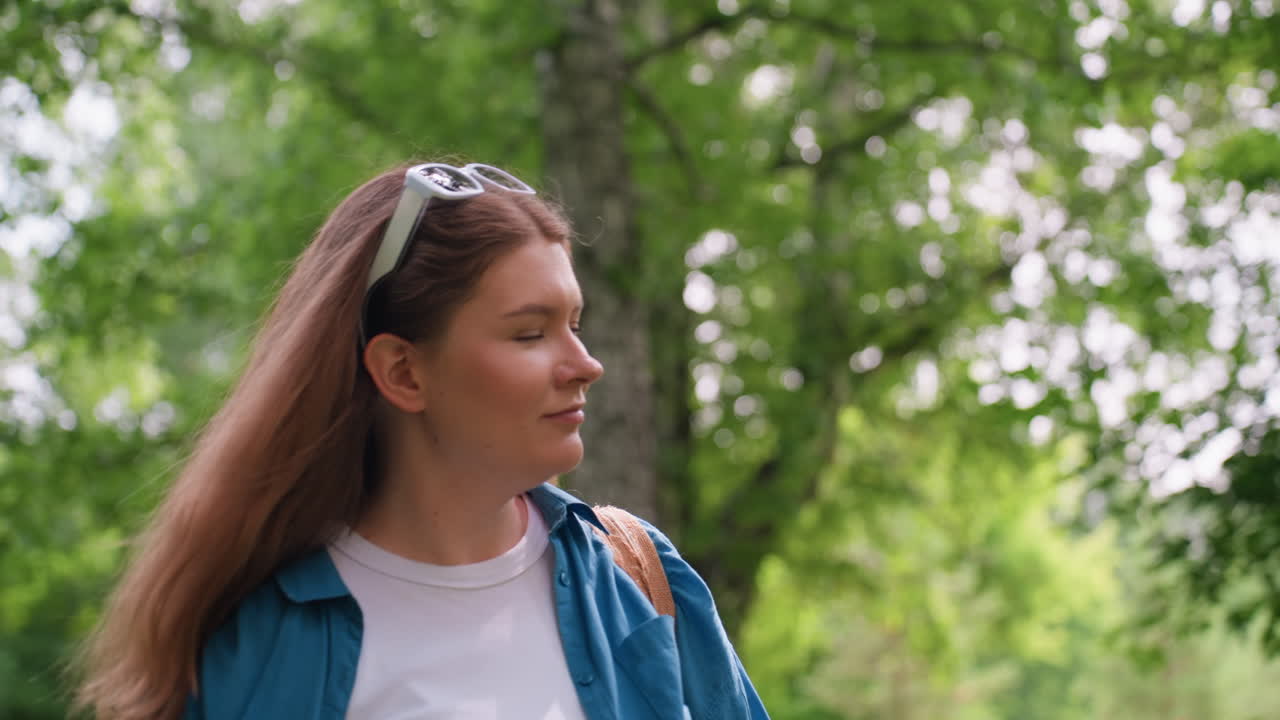 Lady walking through green park admiring nature, wearing blue shirt and white top, looking upward with peaceful smile, surrounded by sunlight and trees, showing calm curiosity