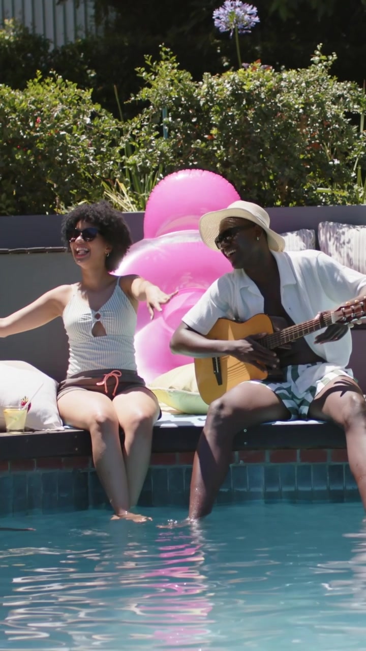 Coppia seduta che indossa costume da bagno e cappello, strimpella la chitarra e saluta a bordo piscina con un galleggiante rosa.
