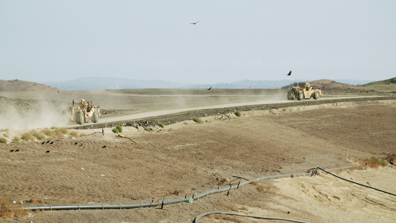 Earthmoving equipment working on a dusty, arid construction site with birds flying overhead and a pipeline in the foreground