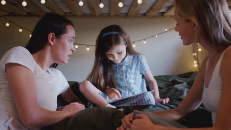 same sex female couple sitting on bed with daughter at home together using digital tablet