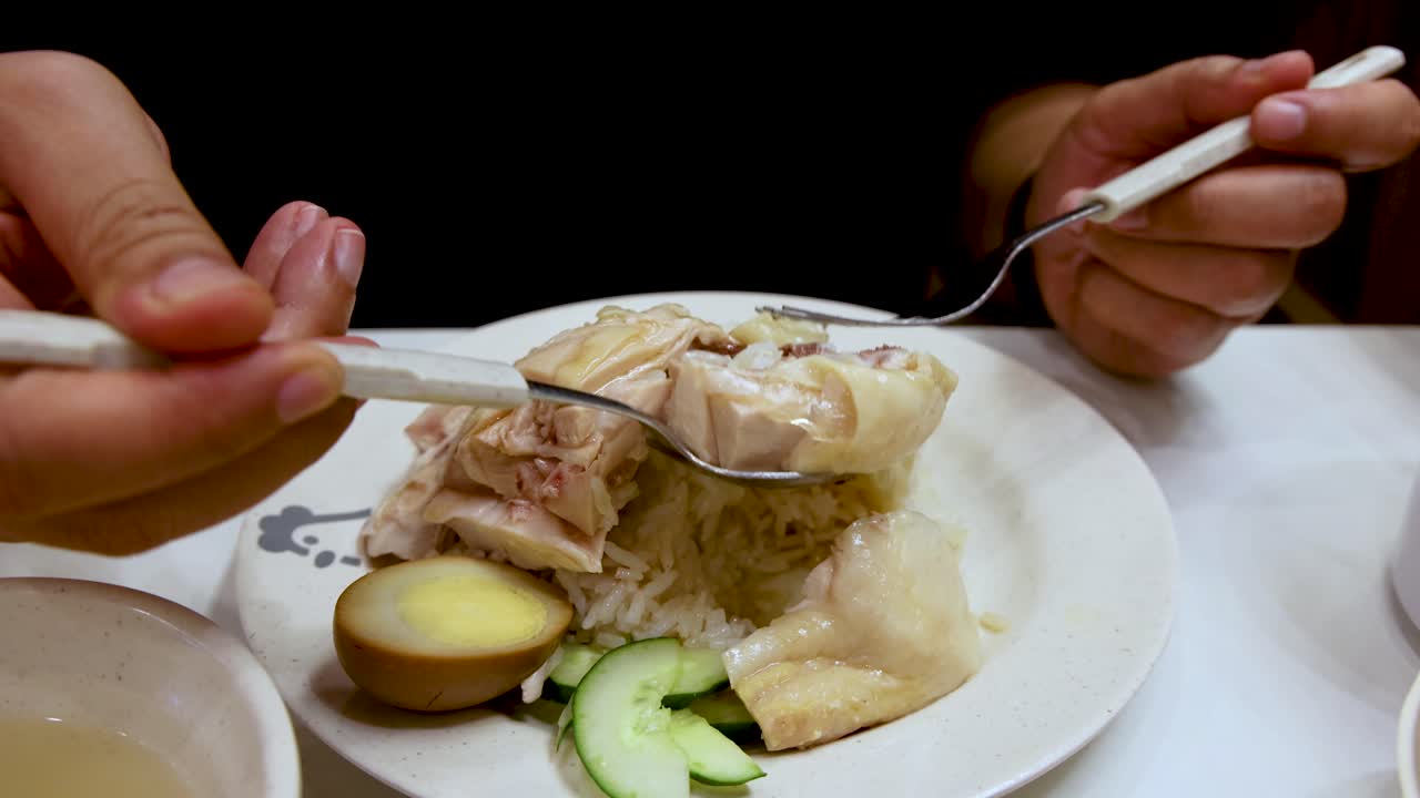 Hands use a spoon and fork to scoop steamed chicken, rice, cucumber, and egg from a white plate under bright indoor lighting, viewed close-up