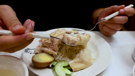 hands use a spoon and fork to scoop steamed chicken, rice, cucumber, and egg from a white plate under bright indoor lighting, viewed close-up