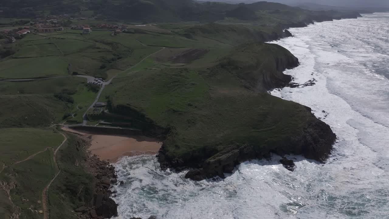 Slow descending drone shot over Santa Tecla beach in Cantabria, Spain, showing green meadows, small coastal villages and rocky cliffs under a partly cloudy summer sky. Peaceful and scenic atmosphere