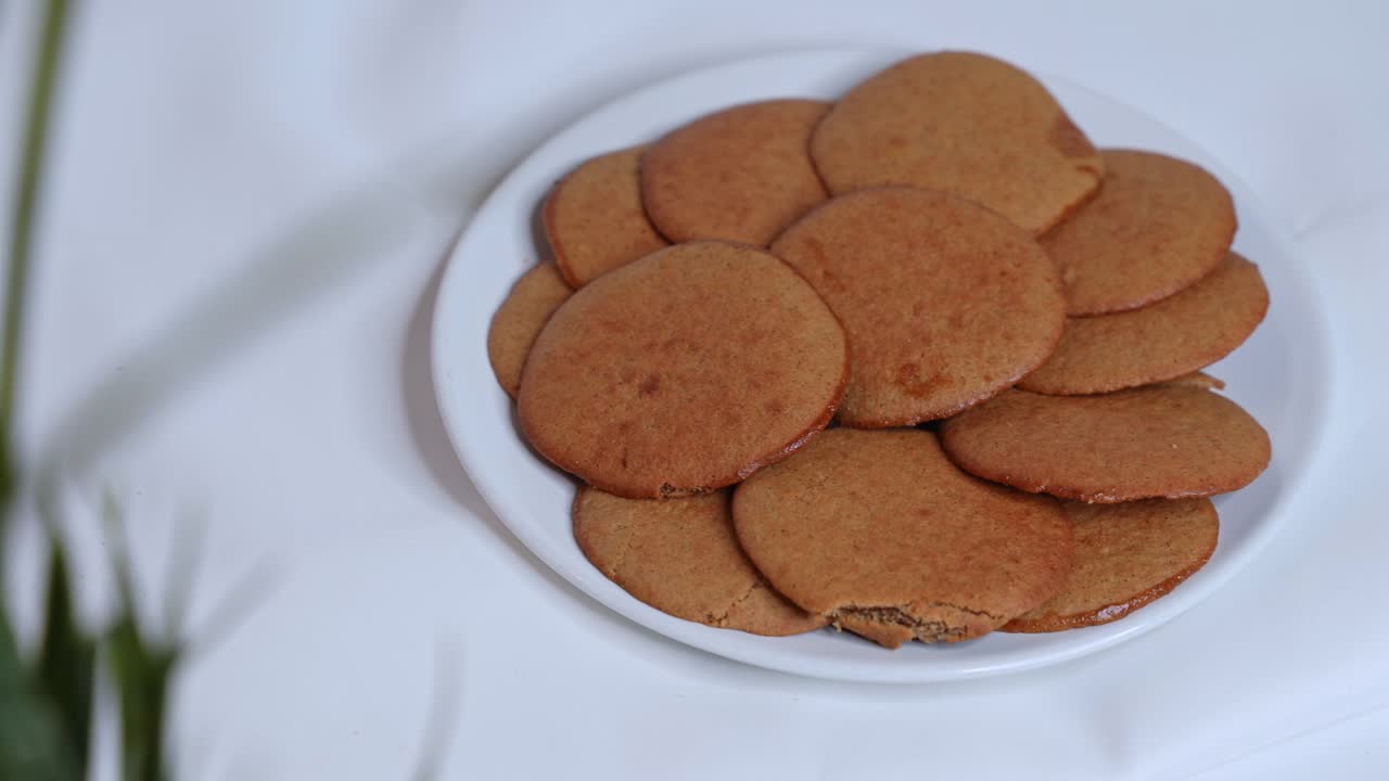 Woman’s hand picks a glistening date-filled cookie from a white plate on a soft white backdrop. Perfect for Middle Eastern food, dessert, or Ramadan-related stock. Shot in stunning 4K detail.