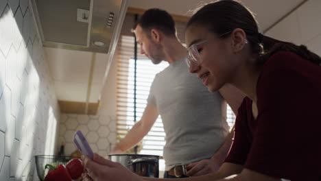 Padre-Caucásico-Con-Hija-Adolescente-Mirando-El-Teléfono-Móvil-En-La-Cocina-Mientras-Cocina