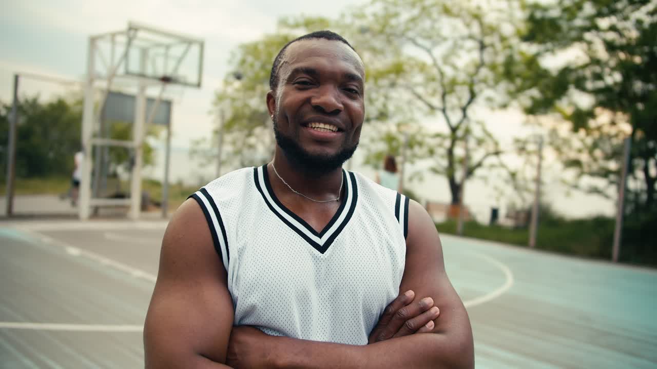 A Black man in a white t-shirt poses in front of his friends who play basketball outdoor on the court in summer