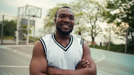 a black man in a white t-shirt poses in front of his friends who play basketball outdoor on the court in summer