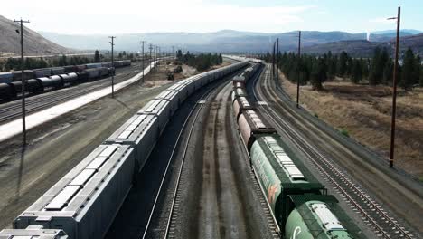 vuelo hacia adelante dolly drone disparado volando entre trenes de carga y trenes tanque en una estación de ferrocarril en un ambiente desértico en un día soleado con montañas en el fondo y líneas eléctricas