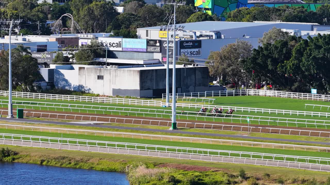 Multiple jockeys ride thoroughbred horses around a sunlit turf racetrack, captured in a wide, panning aerial sequence at Gold Coast, Australia