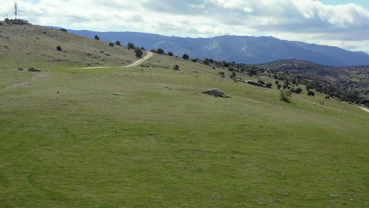vuelo de avión no tripulado en un prado verde con un camino y una cámara ascendente girando descubriendo un fondo increíble de montañas con un cielo con nubes y una colina con una antena en cebreros ávila españa