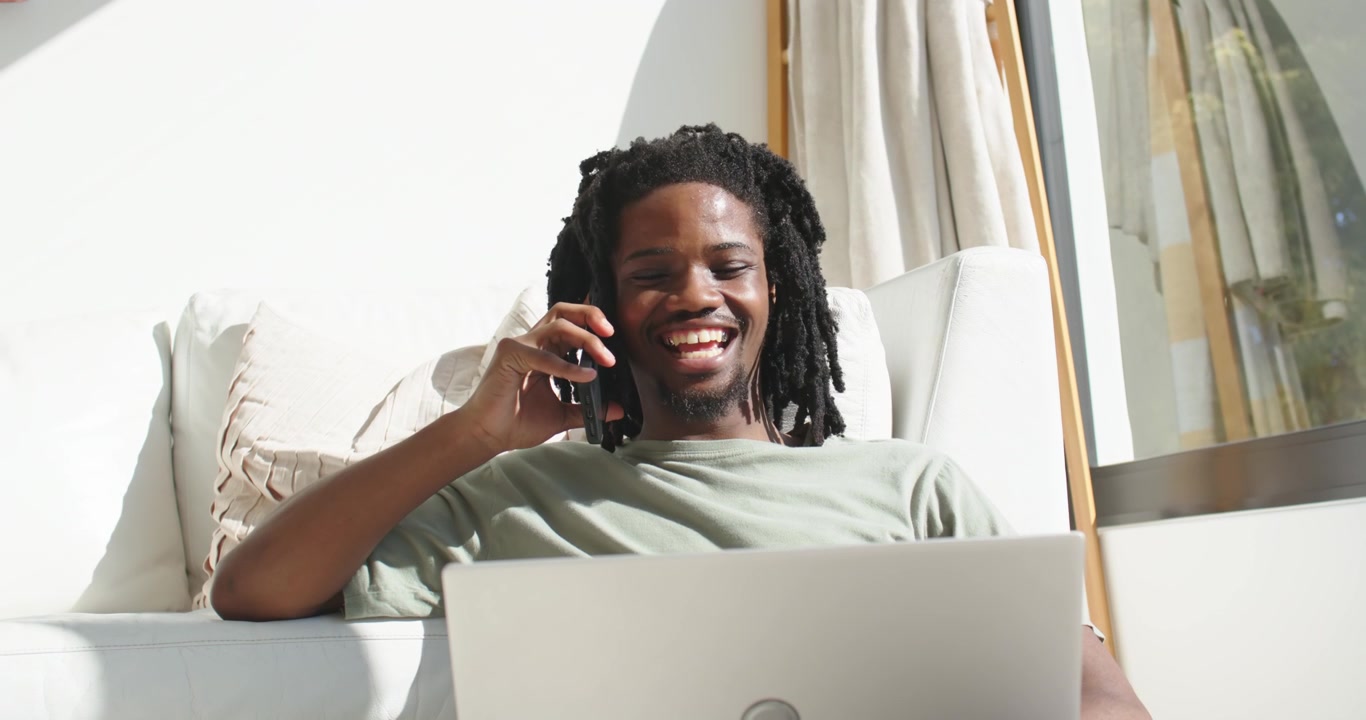 Phone ringing, African American man holding phone, smiling and checking laptop at home for work