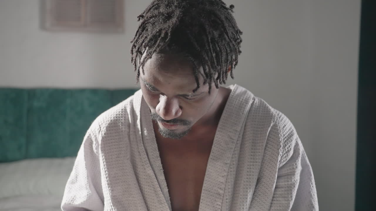 Guy in bathrobe sitting on bed, deeply engrossed in reading a book. Soft light from window enhances peaceful, relaxed indoor atmosphere. Comfortable setting with cozy bedding and tranquil mood