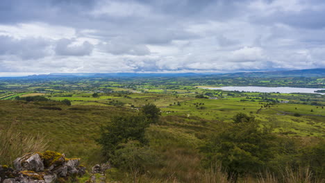 Timelapse-of-rural-nature-farmland-with-rocks,-trees-and-sheep-in-the-foreground-field-and-hills-and-lake-in-distance-during-sunny-cloudy-day-viewed-from-Carrowkeel-in-county-Sligo-in-Ireland