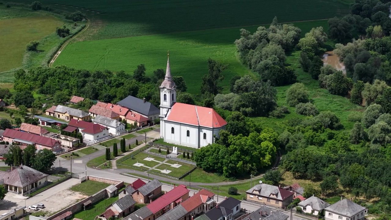 Letecký pohled na malou maďarskou vesnici ukazující její kostel, okolní domy a zemědělská pole pod jasnou oblohou.