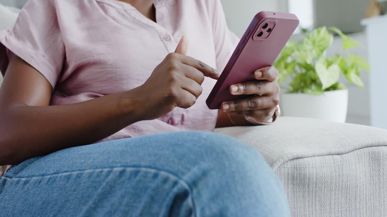 Vertikální video: Žena v růžovém topu a džínách ťuká na telefon s růžovým pouzdrem a posouvá po něm prstem, sedí na pohovce doma