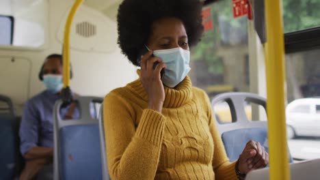 mujer de negocios afroamericana con máscara facial hablando en un teléfono inteligente y sentada en el autobús