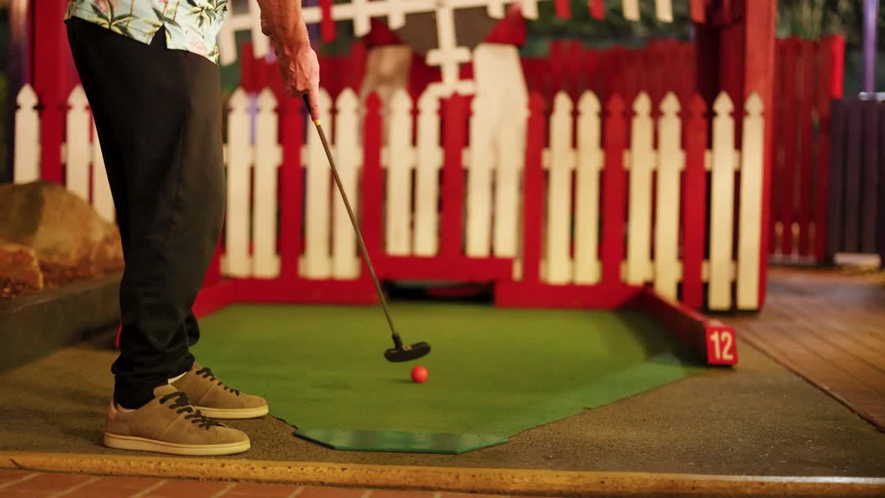 A person putts on a mini-golf course at night, surrounded by red fencing and warm lighting