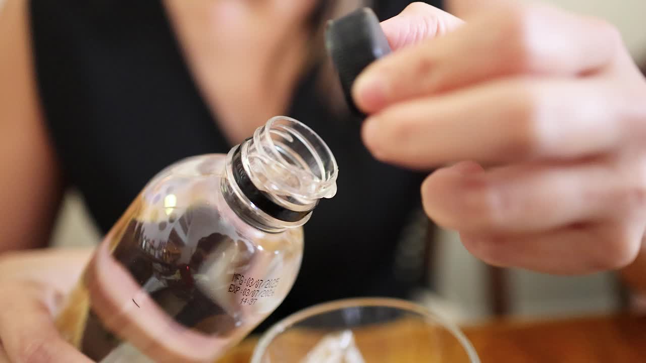 A woman in a sleeveless black top unscrews the cap from a clear glass bottle at a wooden table in soft natural light, close-up perspective