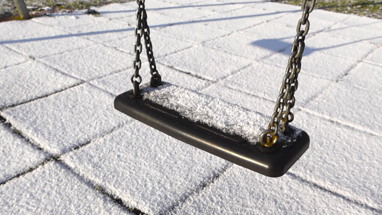 Gliding shot around a swing seat covered in grainy frosty snow. Empty frozen playground under white layer of first snow cover ground. Swing hangs quietly above untouched snow in bright winter sunlight