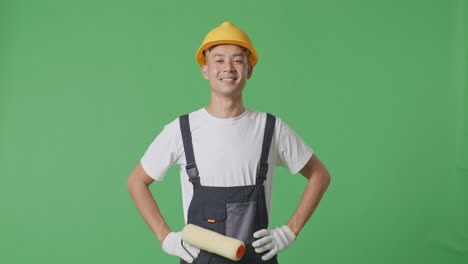asian man painter wearing safety helmet standing with arms akimbo looking at camera and smiling in the green screen background studio