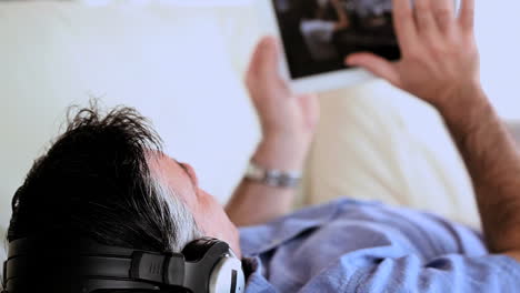 mature man lying on the couch listening to music and using tablet