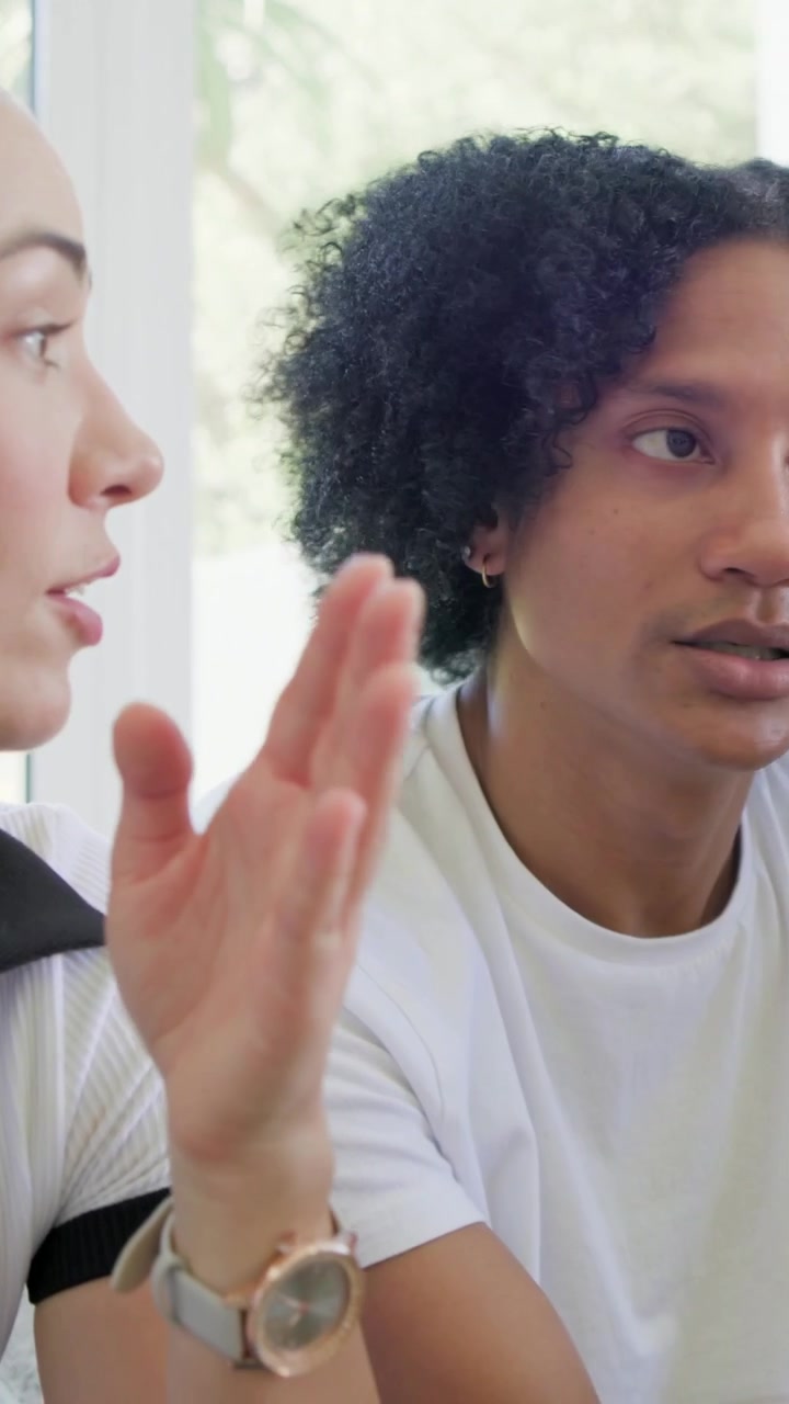 Vertical video: Tilting head woman in white top making point at table by window with green bottle