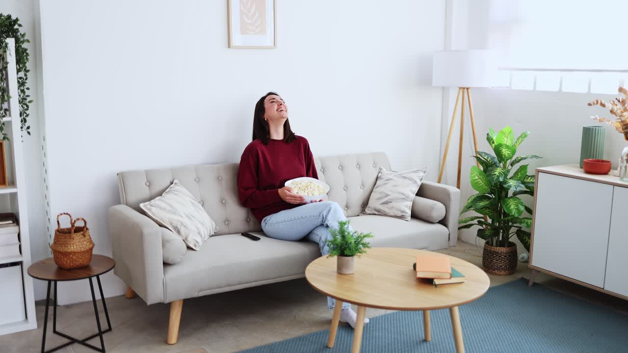 Giovane donna felice che ride mentre guarda la TV e mangia popcorn sul divano