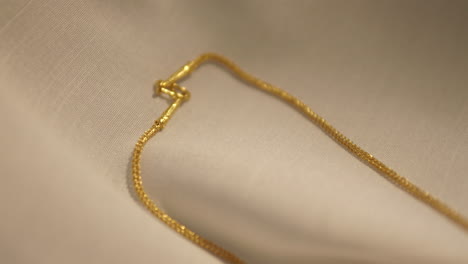 panning from the upper left to the bottom right of the frame over the gold chained necklace that is placed on a cream-colored cloth