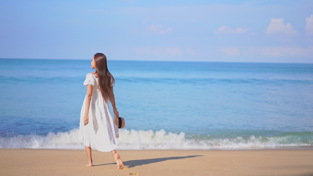 흰색 sundress를 입고 밀짚 모자를 들고 여름에 물가 해변에 서있는 아시아 여자
