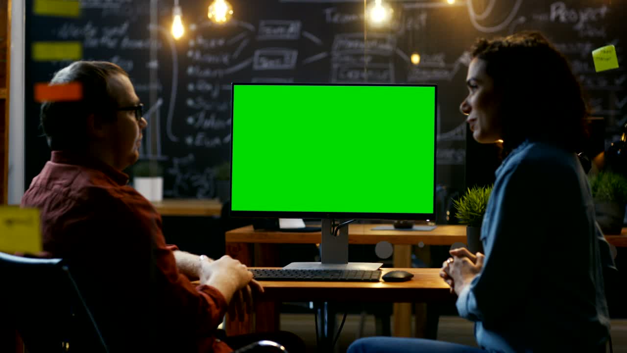 Beautiful Female Office Employee at Her Desk Talks with Her Male Coworker on the Desk Stands Computer with Mock-up Green Screen. Creative Evening Office.