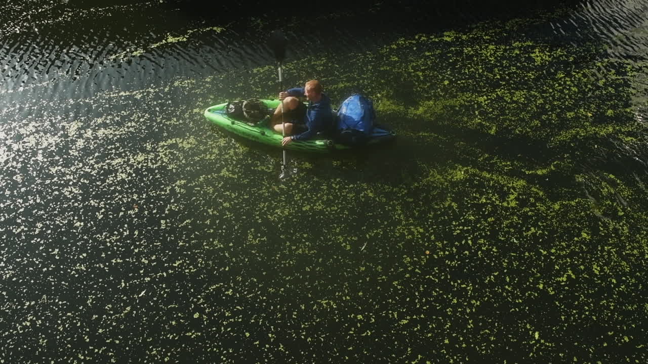 A high angle wide static shot of a river near London with an adult male paddling through on a lime green kayak peacefully